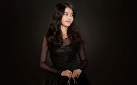 IU, a renowned Asian singer, is pictured in an HD desktop wallpaper. She wears a black dress, standing against a dark background, with a gentle smile on her face.