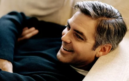 HD PC desktop wallpaper featuring celebrity George Clooney smiling while reclining on a beige sofa.