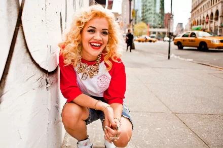A vibrant HD desktop wallpaper featuring Rita Ora laughing and posing playfully in a city setting, showcasing her unique style and charisma.