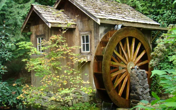 HD desktop wallpaper featuring a rustic man-made watermill nestled in lush green foliage, showcasing weathered wood and a large wooden water wheel.