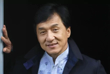 HD wallpaper of celebrity Jackie Chan, featuring a close-up shot of him smiling in a dark jacket and white shirt.