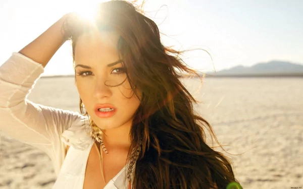 Demi Lovato poses in a sunlit desert landscape, her hair flowing freely. This HD desktop wallpaper captures her striking beauty and vibrant energy against a serene backdrop.