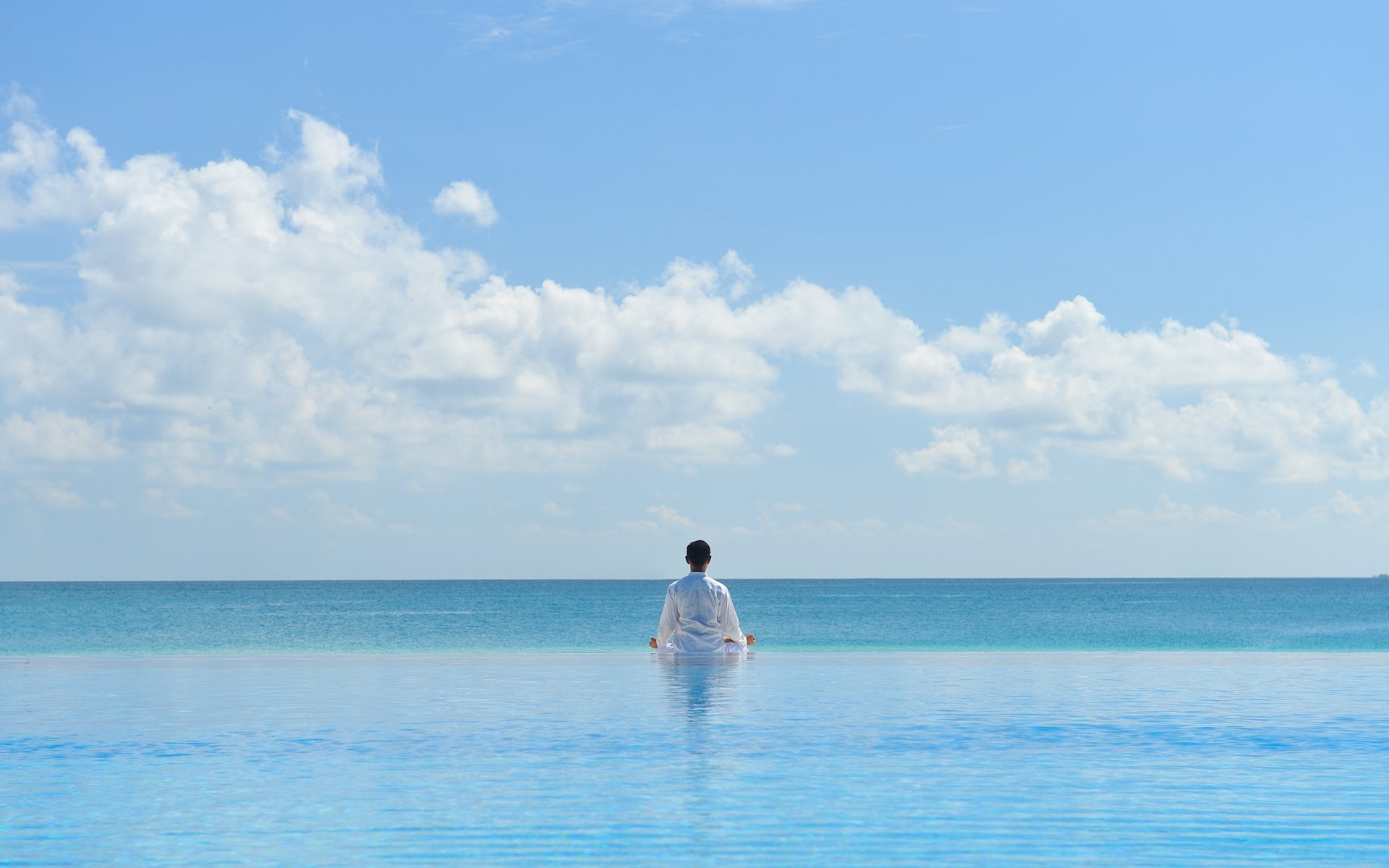 HD desktop wallpaper featuring a person in white sitting peacefully by a tranquil blue pool, facing the calm ocean under a bright sky, evoking a serene religious and zen atmosphere.