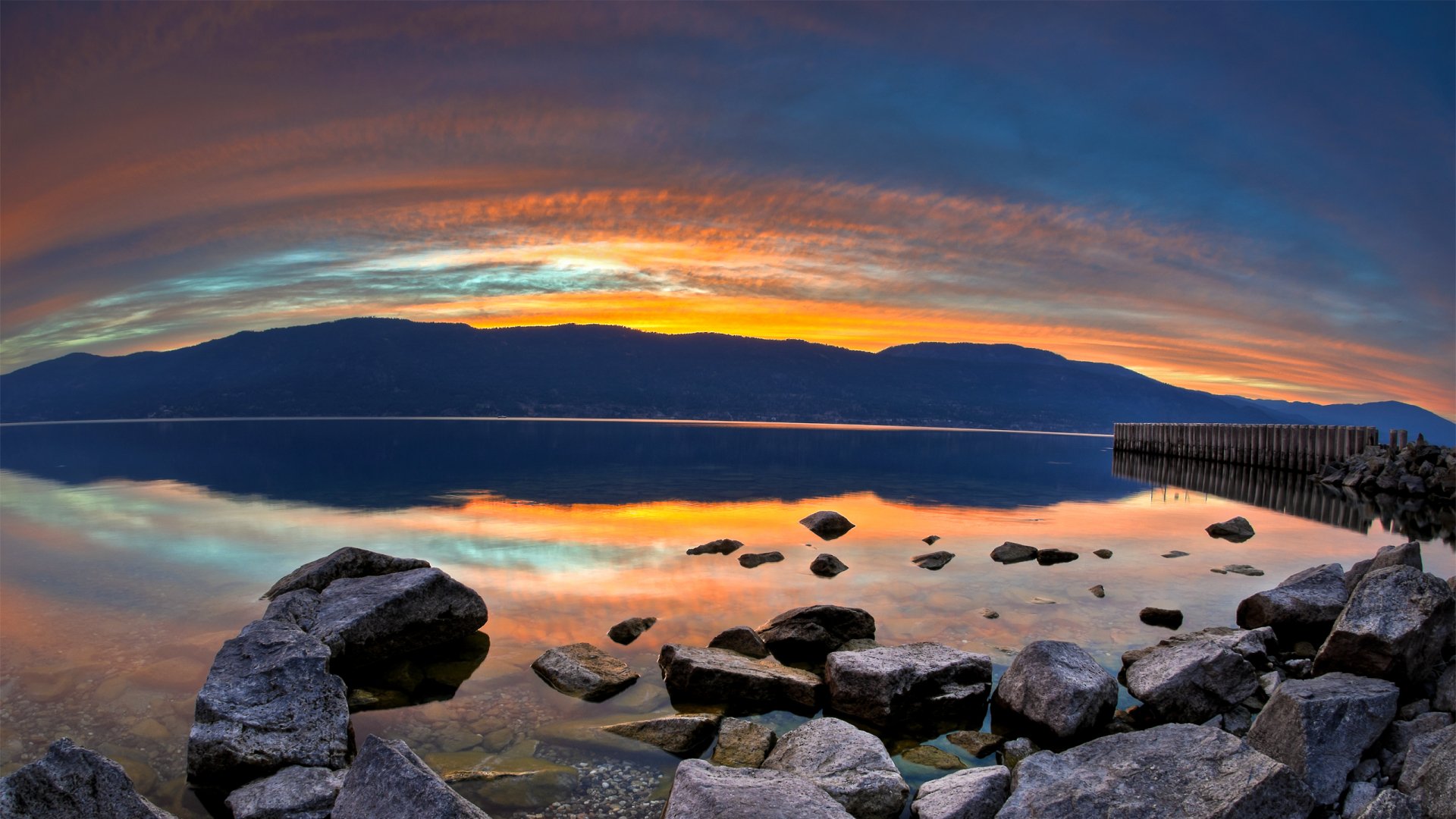 Nature sunrise over a calm lake with rocky shore and distant mountains, vibrant banded sky reflected in water — 2K Quad HD PC desktop wallpaper and background.