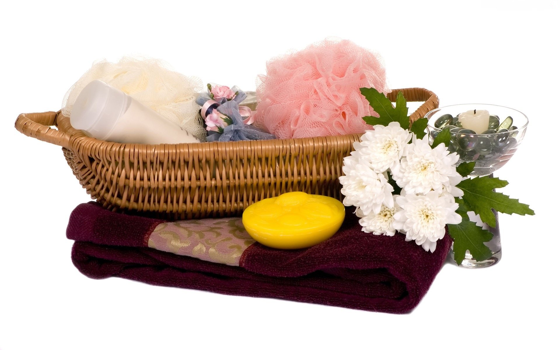 A still life HD desktop wallpaper featuring a wicker basket with spa items, soft towels, a yellow soap, pink bath puff, white flowers, and a small glass bowl with water.