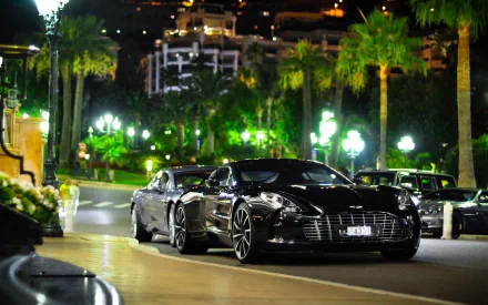 An Aston Martin One-77 parked on a palm-lined street at night, illuminated by soft lights, showcasing its sleek design against an elegant urban backdrop.
