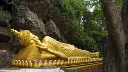 A serene image of a golden reclining Buddha statue nestled against a rocky backdrop, surrounded by lush greenery, capturing the essence of Buddhism in a tranquil setting.