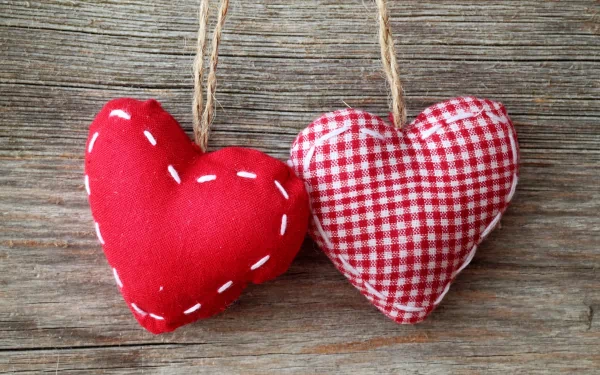 Artistic HD desktop wallpaper featuring two hanging fabric hearts—one solid red with white stitches, the other red and white checkered—against a rustic wooden background.