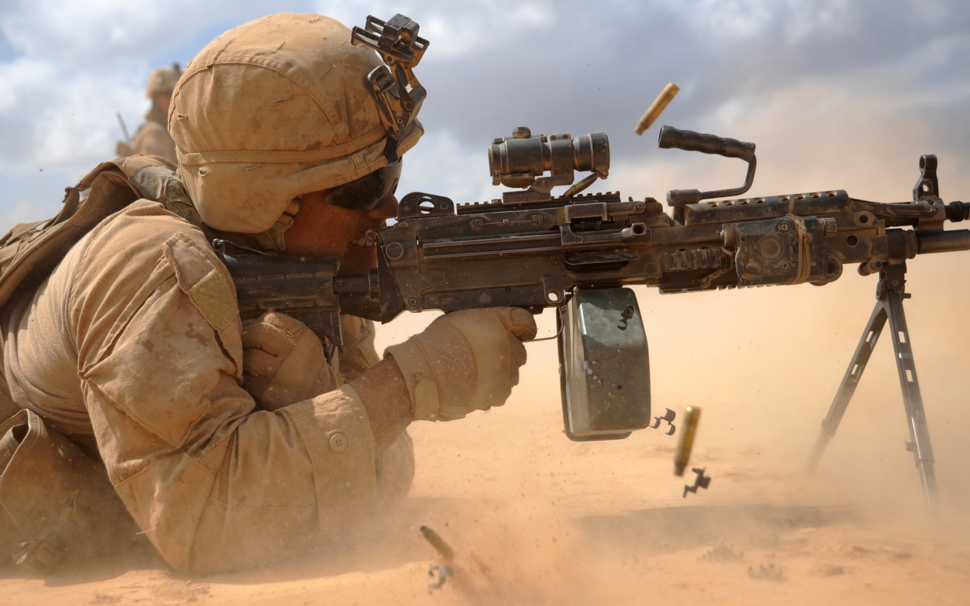 A soldier in military gear aims an M249 SAW in a sandy environment, with spent cartridges flying, capturing the intensity of combat in a high-definition wallpaper.