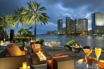 A serene terrace in Miami features lush palm trees and elegant seating, overlooking a skyline of skyscrapers against a twilight sky, with drinks and candles enhancing the atmosphere.