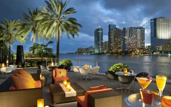 A serene terrace in Miami features lush palm trees and elegant seating, overlooking a skyline of skyscrapers against a twilight sky, with drinks and candles enhancing the atmosphere.