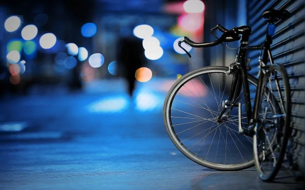 A bicycle leans against a building on a dimly lit city street at night, with colorful bokeh lights in the background, evoking a mood of solitude. HD desktop wallpaper.