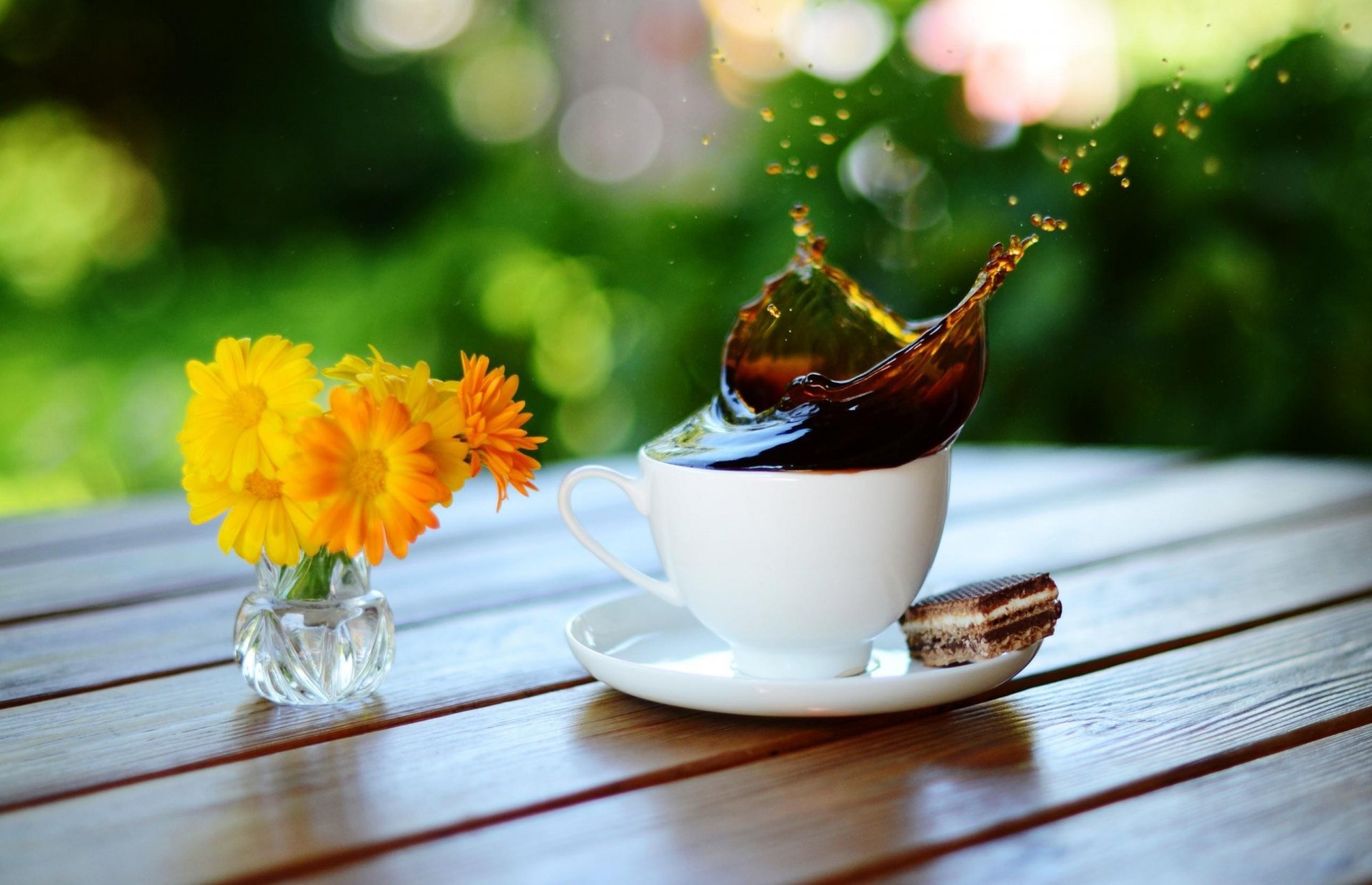 HD desktop wallpaper featuring a white coffee cup on a wooden table with coffee splashing out, next to a small vase of vibrant yellow flowers, set against a blurred green background.