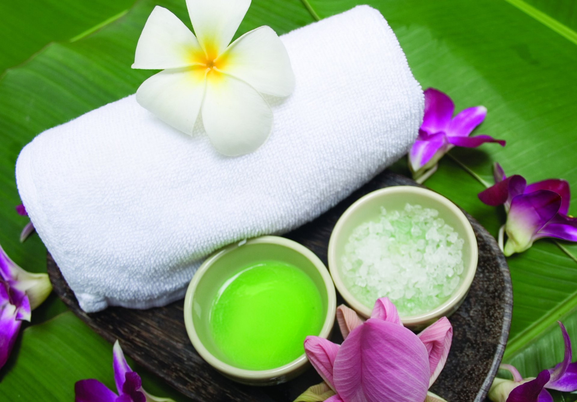 2K Quad HD PC desktop wallpaper of a man-made spa arrangement: white towel, bowls of green oil and bath salts, frangipani and orchid flowers on a banana leaf.