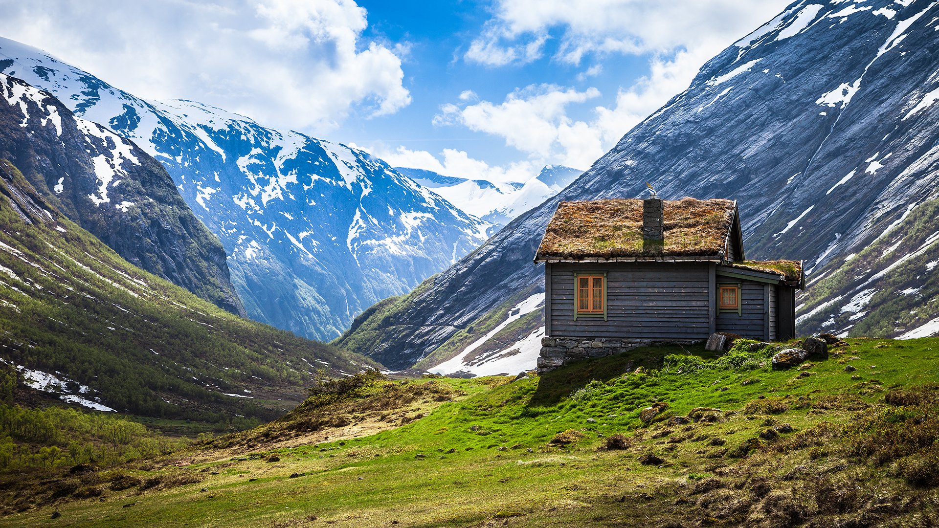 HD desktop wallpaper of a man-made cabin with a grass roof, set on a green hillside surrounded by snow-capped mountains under a bright blue sky.