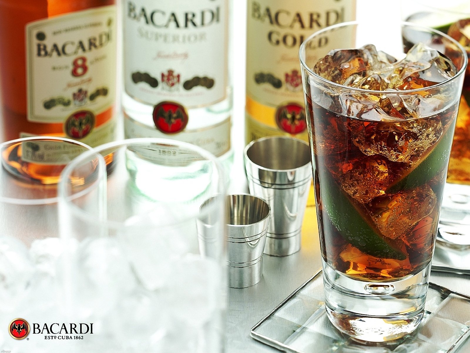 HD desktop wallpaper showing a close-up of a glass filled with a dark liquor drink and ice, surrounded by Bacardi liquor bottles and metal measuring jiggers on a reflective surface.