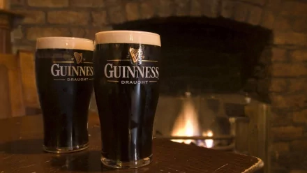 Two glasses of Guinness Draught sit on a table in front of a warm fireplace, creating a cozy atmosphere. This HD wallpaper captures a inviting moment for beer enthusiasts.