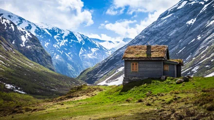 HD desktop wallpaper of a man-made cabin with a grass roof, set on a green hillside surrounded by snow-capped mountains under a bright blue sky.
