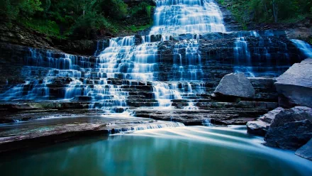 Albion Falls, a stunning waterfall in Canada, cascades over rocky ledges into a serene pool, surrounded by lush greenery in this HD nature-themed desktop wallpaper.