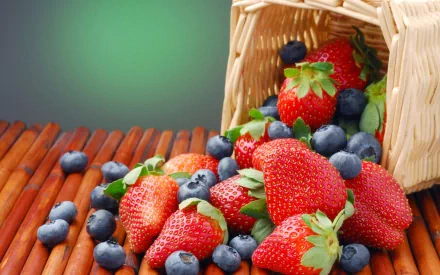 HD desktop wallpaper featuring a close-up of fresh strawberries and blueberries spilling out of a wicker basket on a wooden surface with a soft green background.