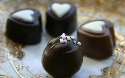 HD desktop wallpaper featuring an elegant close-up of assorted chocolates on an ornate gold and white plate. The chocolates are adorned with delicate white hearts and decorative toppings.