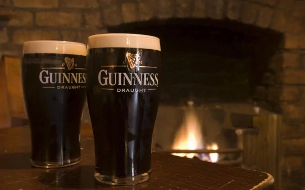 Two glasses of Guinness Draught sit on a table in front of a warm fireplace, creating a cozy atmosphere. This HD wallpaper captures a inviting moment for beer enthusiasts.