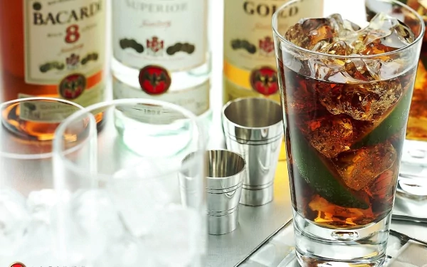 HD desktop wallpaper showing a close-up of a glass filled with a dark liquor drink and ice, surrounded by Bacardi liquor bottles and metal measuring jiggers on a reflective surface.