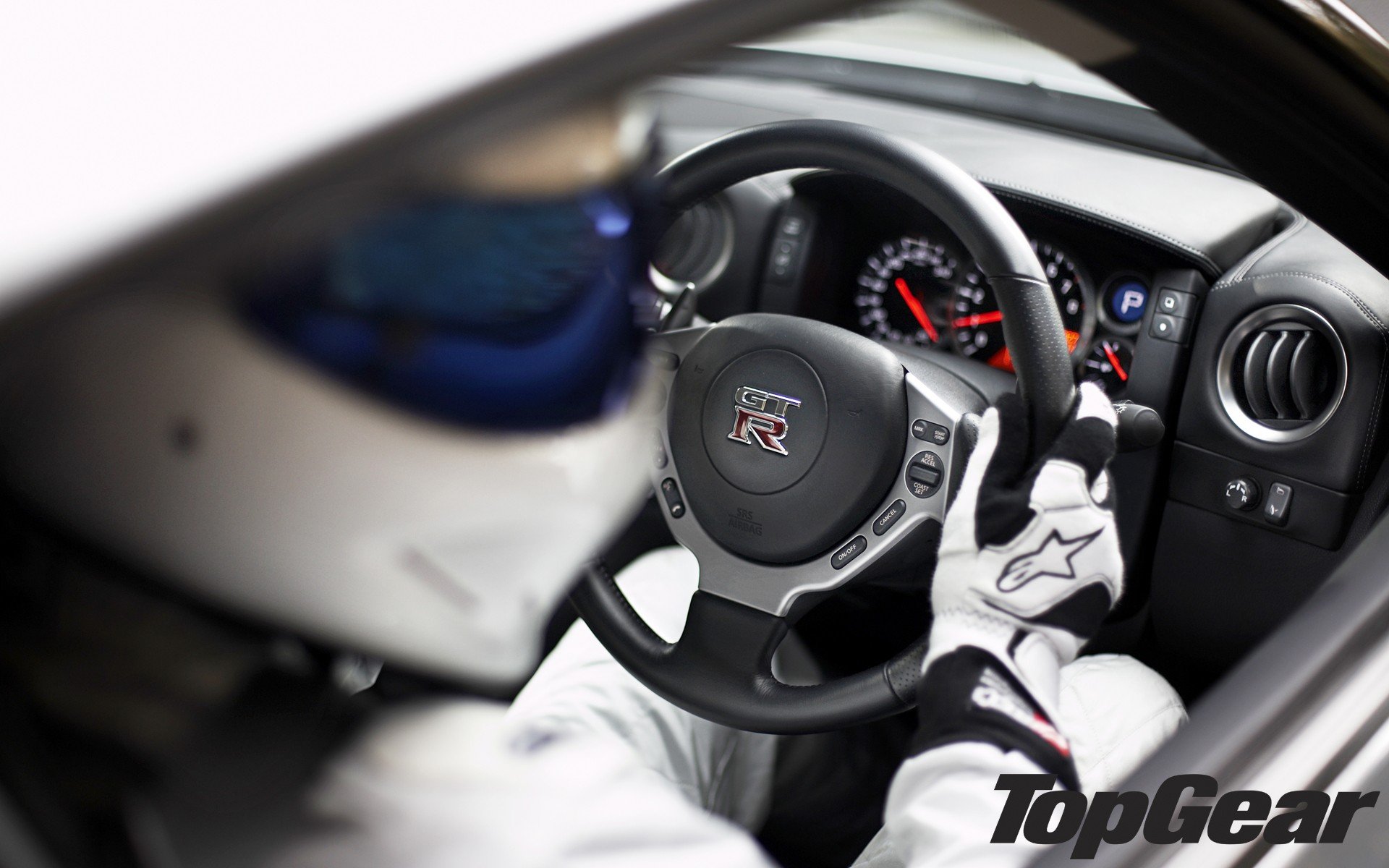 HD desktop wallpaper of a Top Gear-themed steering wheel and dashboard view, with a blurred driving suit in the background.