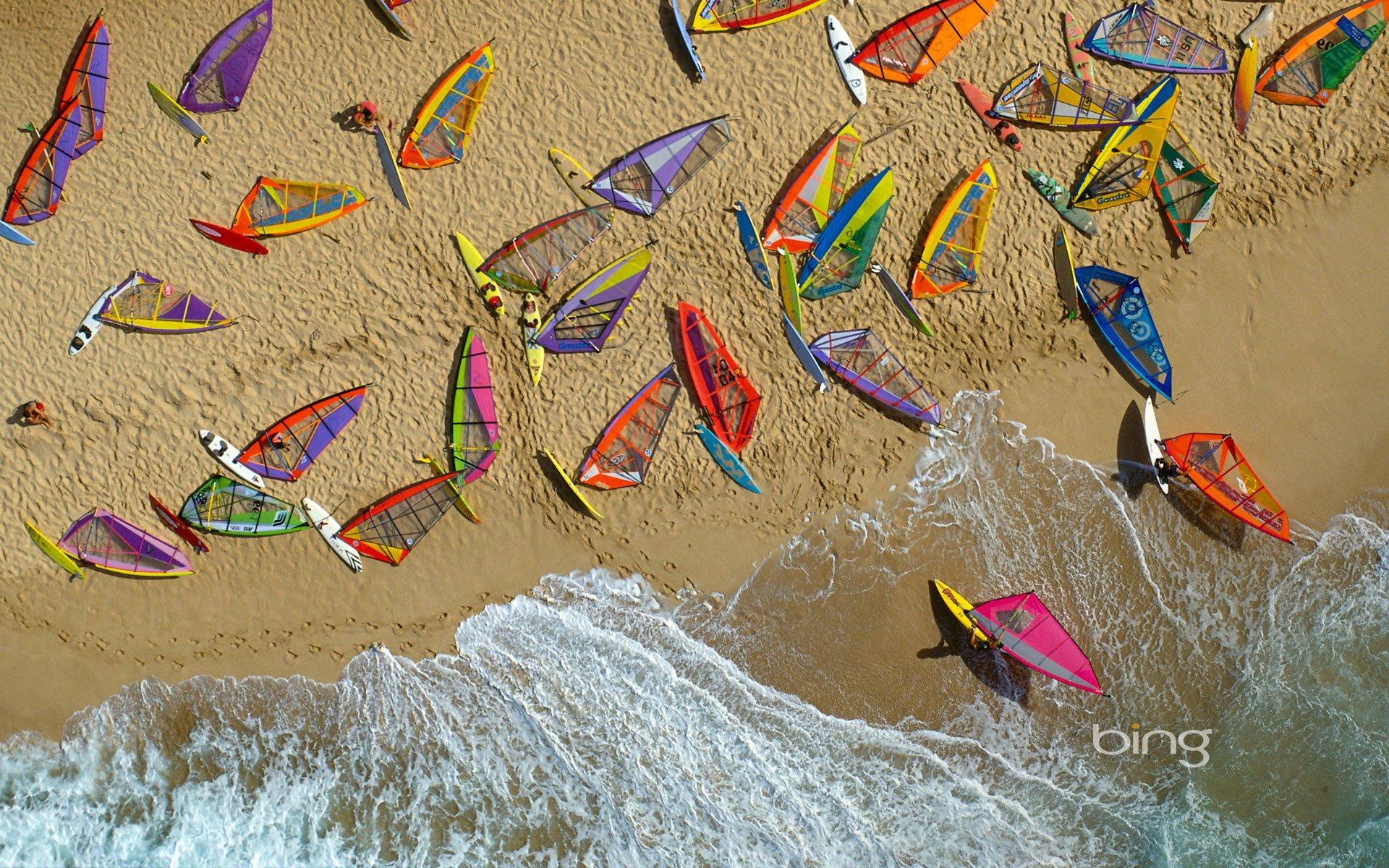 HD PC desktop wallpaper and background: aerial view of colorful windsurfing sails and boards scattered on a sandy beach as waves lap the shore — Sports.