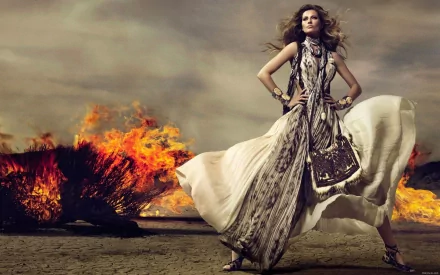 HD desktop wallpaper featuring celebrity Gisele Bündchen in a flowing dress, standing confidently with flames blazing in the background under a dramatic sky.