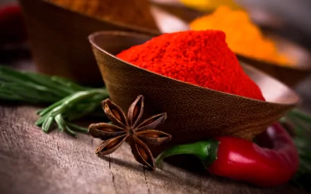 HD desktop wallpaper showing vibrant herbs and spices including bright red chili powder, star anise, green chili, and fresh herbs on a rustic wooden surface.