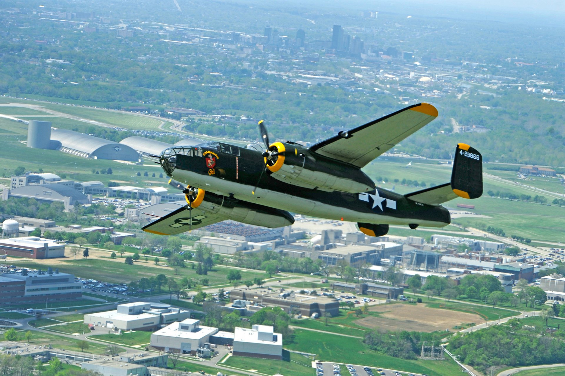 HD PC desktop wallpaper featuring a vintage military aircraft flying over a cityscape on a clear day.