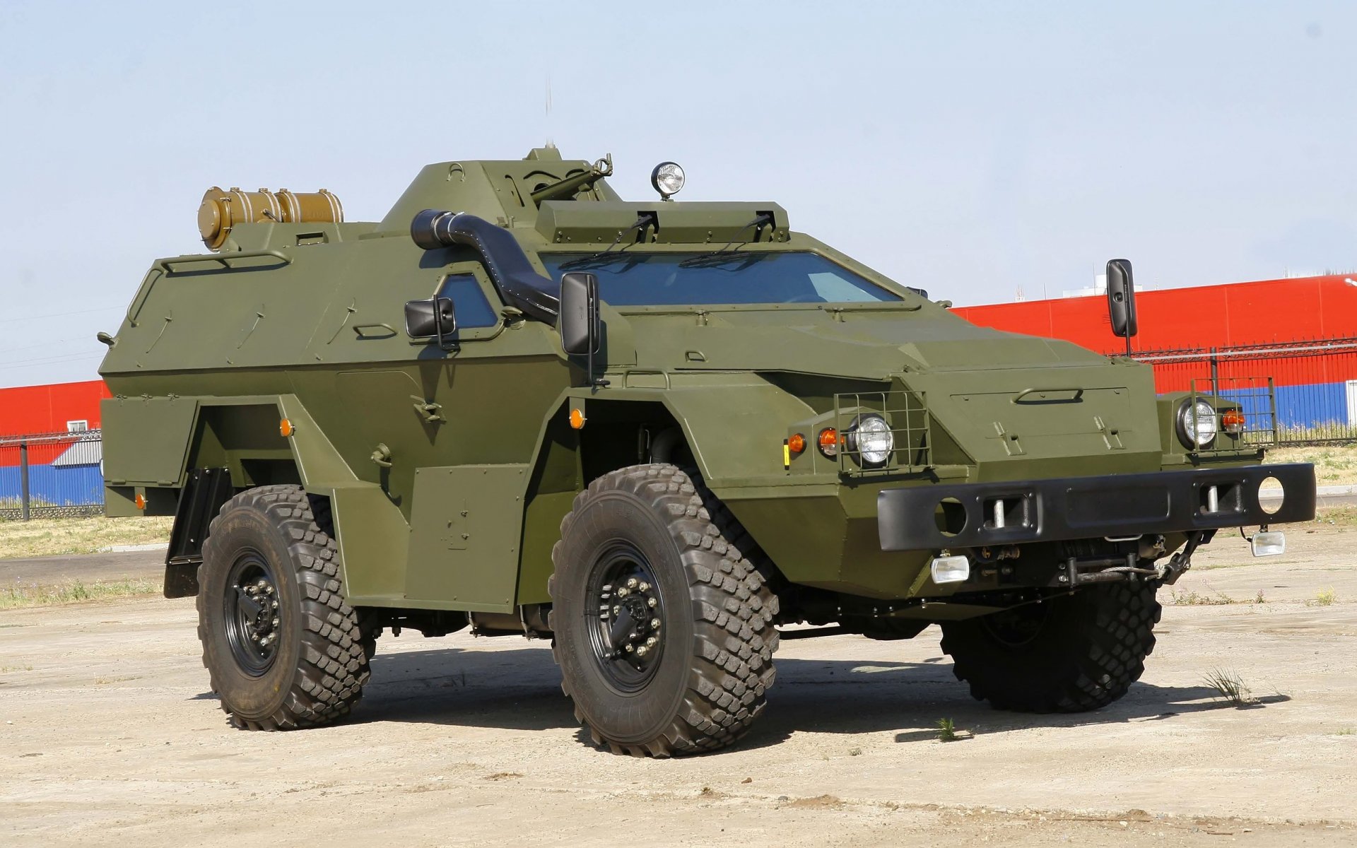 2K Quad HD PC desktop wallpaper: olive-green armored military vehicle parked on concrete with clear sky and red-blue industrial backdrop