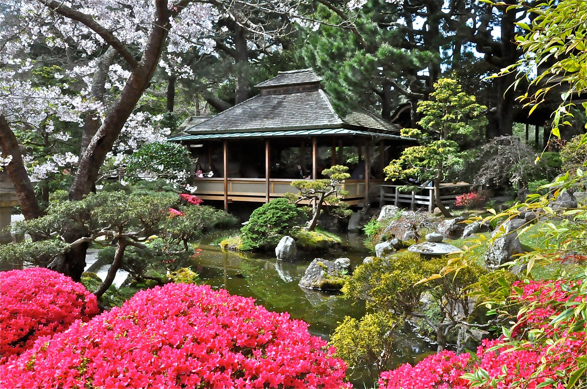 HD PC desktop wallpaper showing a serene man-made garden with a traditional house surrounded by vibrant flowers, trees, and a calm pond.