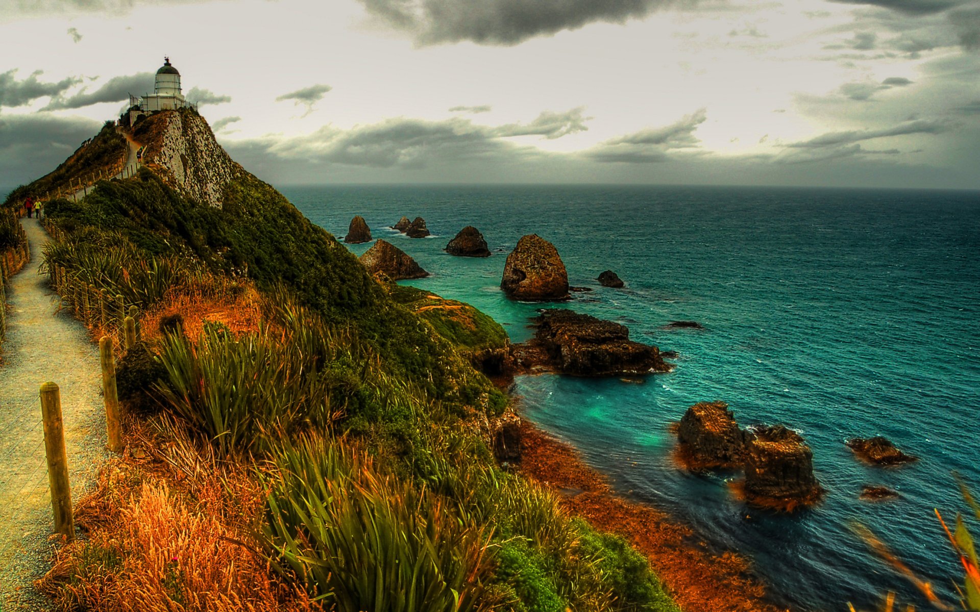 Photography of a rugged coastline with a cliffside lighthouse, winding path and rocky islets over teal sea under dramatic skies — HD PC Desktop Wallpaper and Background