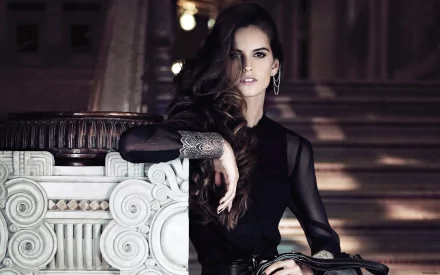 Portrait of woman Izabel Goulart wearing a black outfit, posing confidently by an ornate white column with dimly lit stairs in the background, HD desktop wallpaper.