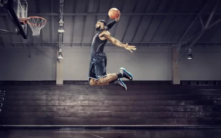 HD wallpaper of a basketball player in mid-air, poised to dunk, showcasing athleticism and intensity in a sports setting. The dimly lit indoor court adds drama to the scene.