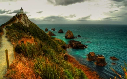 Photography of a rugged coastline with a cliffside lighthouse, winding path and rocky islets over teal sea under dramatic skies — HD PC Desktop Wallpaper and Background