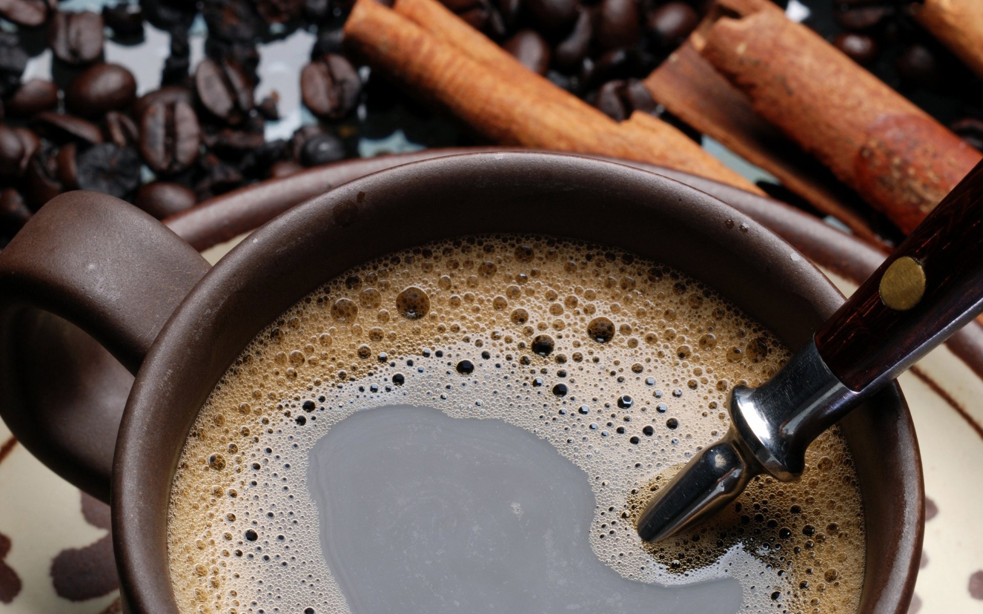 Close-up of frothy coffee in a brown mug with a spoon, cinnamon sticks and scattered beans — 2K Quad HD PC desktop wallpaper and background (food, coffee).
