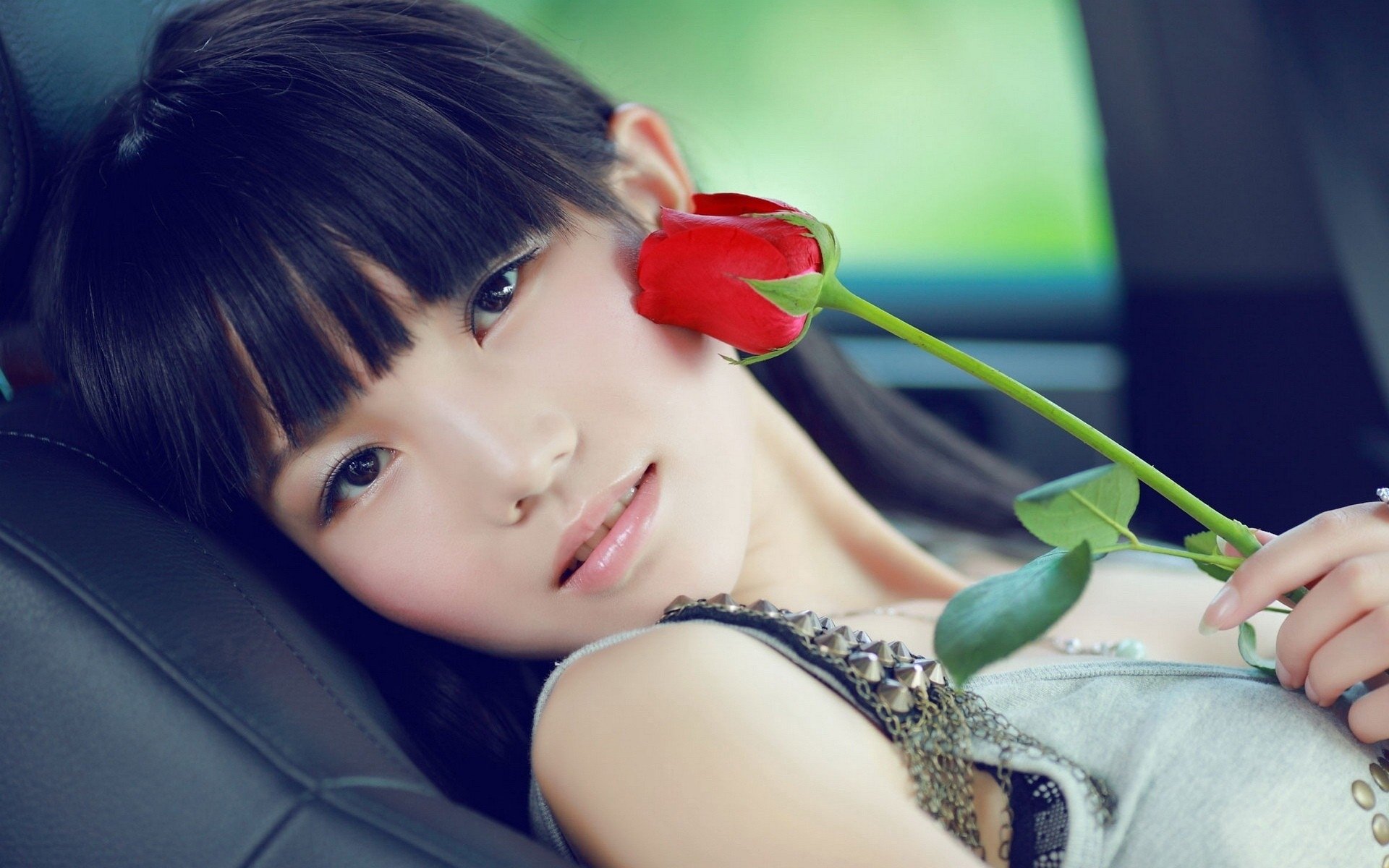 HD wallpaper featuring an Asian woman lying on a car seat, holding a red rose to her cheek. Background showcases a soft green blur, enhancing the serene and intimate mood.
