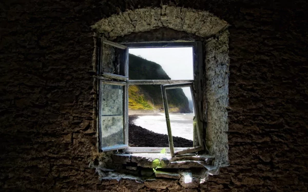 HD PC desktop wallpaper: view through a man-made stone window with open panes onto a sunlit rocky coastline and sea, a small plant growing on the weathered sill.