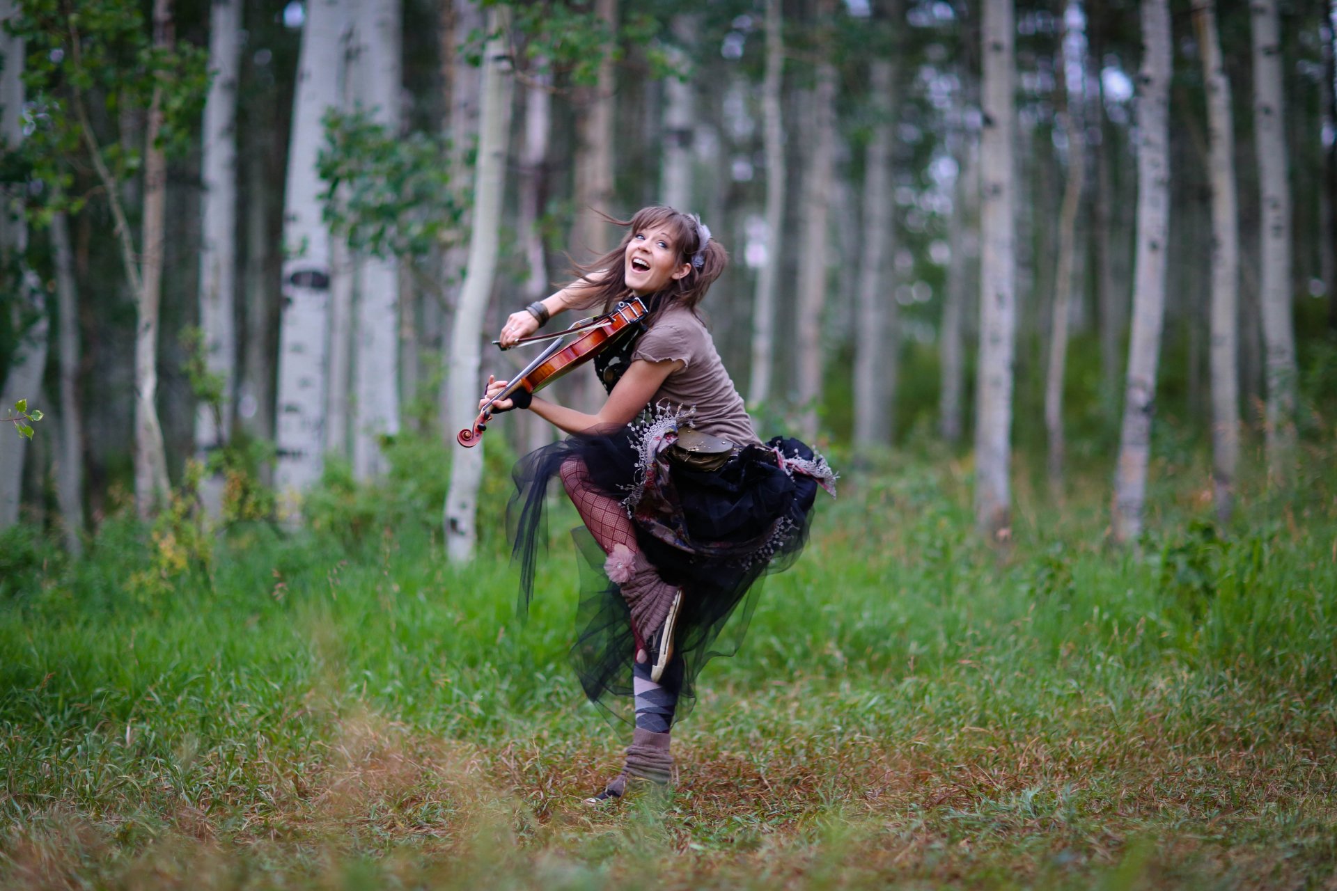 A joyful musician, Lindsey Stirling, plays the violin amid a lush forest backdrop, embodying a vibrant spirit in this 4K Ultra HD desktop wallpaper.