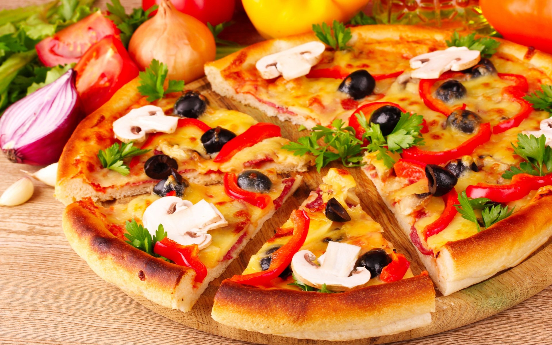 HD desktop wallpaper featuring a delicious pizza topped with mushrooms, olives, tomatoes, onions, and parsley, surrounded by fresh ingredients on a wooden surface.