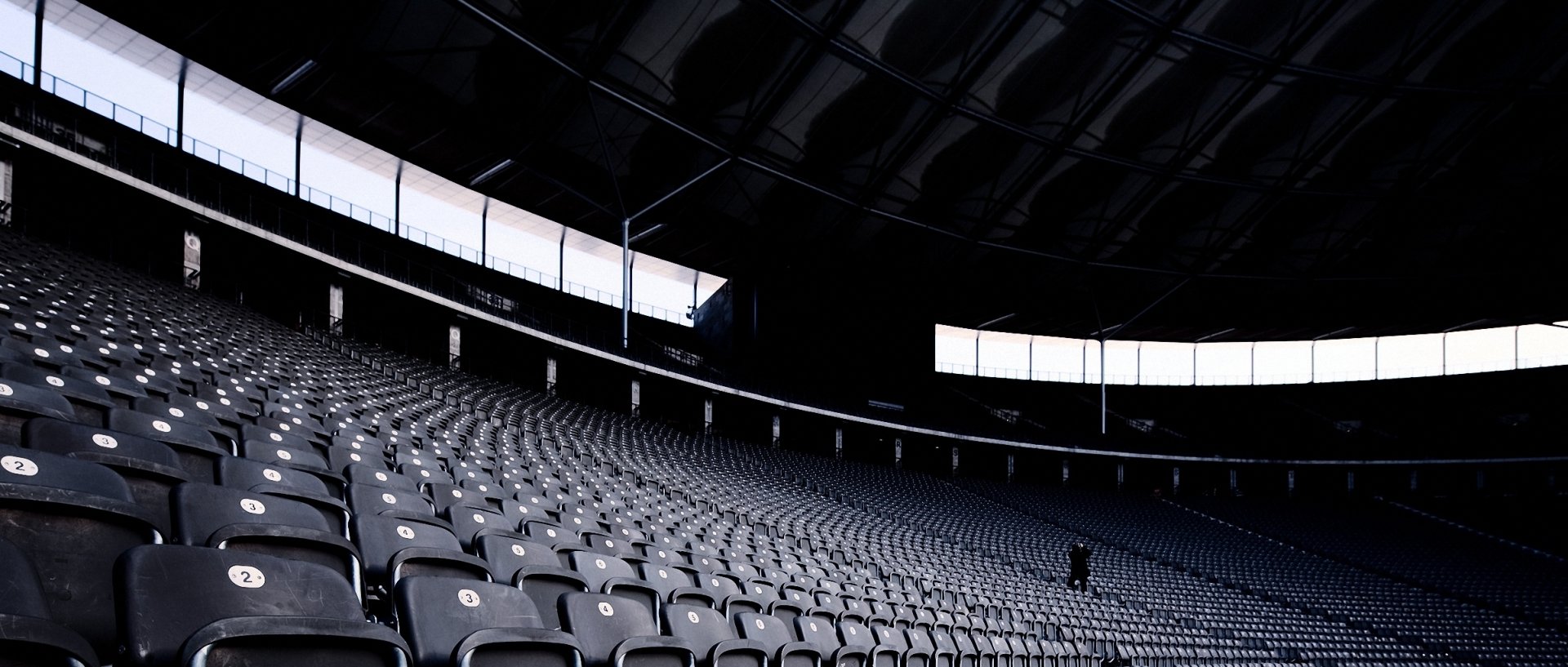 HD desktop wallpaper of a vast, dimly lit sports stadium interior with rows of empty seats curving toward the illuminated roof and a solitary figure in the distance.