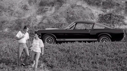 Black-and-white HD PC desktop wallpaper of a vehicle: 1966 Ford Mustang GT350H parked by a hillside, with a couple walking in the grassy foreground.