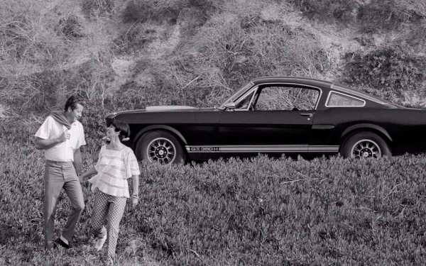 Black-and-white HD PC desktop wallpaper of a vehicle: 1966 Ford Mustang GT350H parked by a hillside, with a couple walking in the grassy foreground.