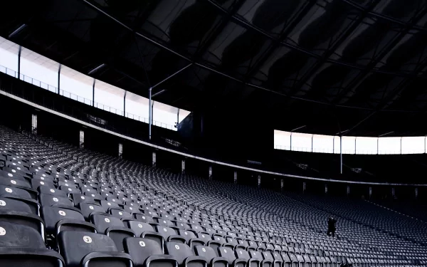 HD desktop wallpaper of a vast, dimly lit sports stadium interior with rows of empty seats curving toward the illuminated roof and a solitary figure in the distance.