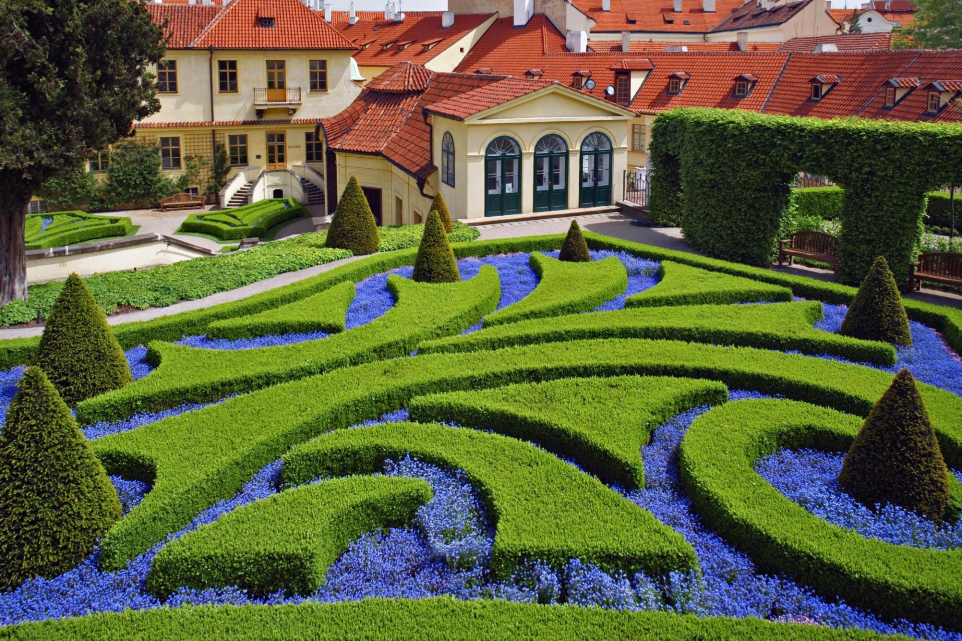 HD desktop wallpaper featuring Prague Botanical Garden in the Czech Republic, showcasing manicured hedges and vibrant flower beds against charming historic buildings.