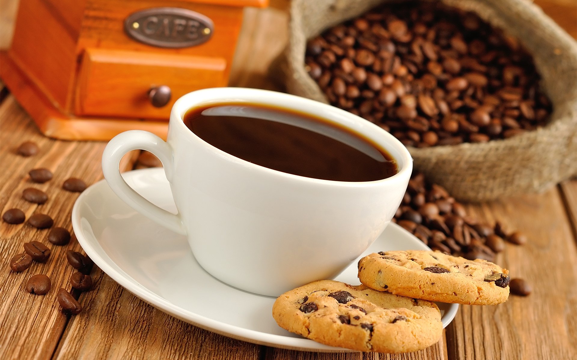 HD desktop wallpaper featuring a white cup of black coffee on a saucer with two chocolate chip cookies, coffee beans scattered on a wooden surface, and a vintage grinder in the background.