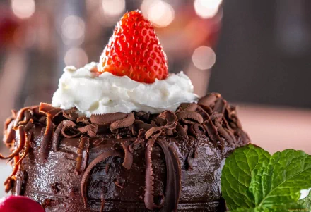 A close-up of a decadent chocolate cake topped with whipped cream and a fresh strawberry, adorned with chocolate shavings, creating an inviting wallpaper for food lovers.